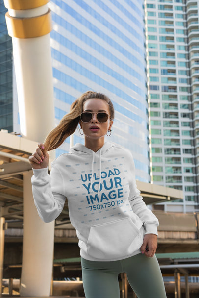Pullover Hoodie Mockup Featuring a Woman Posing in Front of Some Tall Buildings